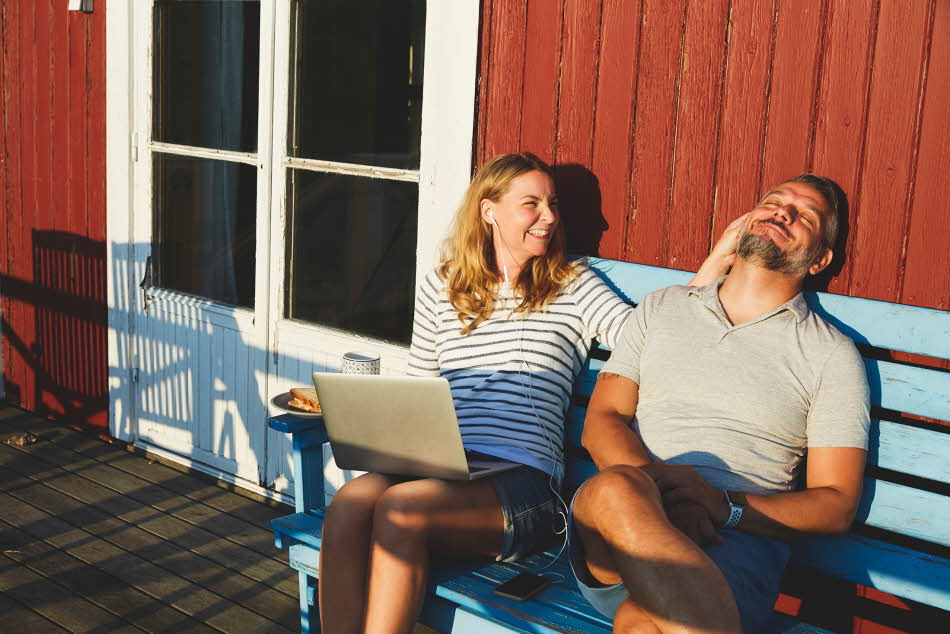 Mann med grå t-skjorte med v-hals sitter sammen med dame på blå benk i hytteveggen