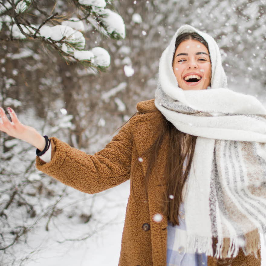 Dame med brun teddykåpe og hvitt skjerf tullet rundt hodet smiler til kamera mens hun kaster snø i været.