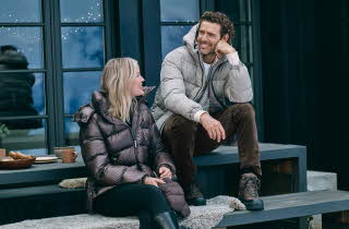 En mann i beige boblejakke og en kvinne i brun boblejakke sitter ute på et bord.