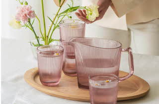 Et fat i tre med en rosa mugge og fire rosa glass, mens en person ordner blomster i en vase i bakgrunnen