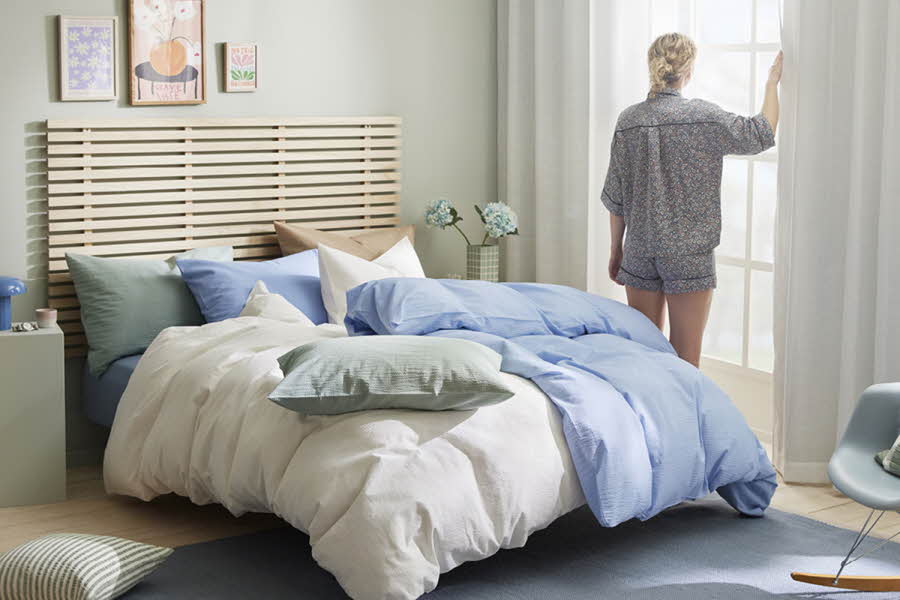 Et soverom, hvor en kvinne står ved et vindu og åpner gardinene. Sengen er redd opp i blått, grønt og hvitt