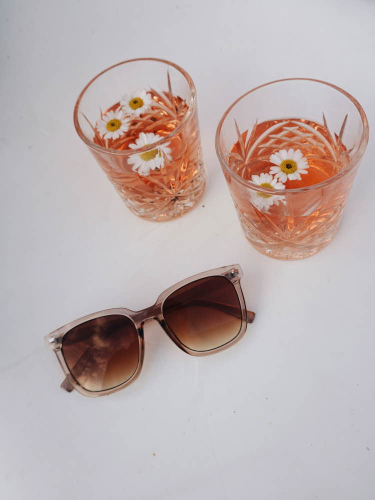 To glass med oransj drikke og hvit blomsterpynt, samt et par solbriller på et hvitt bord