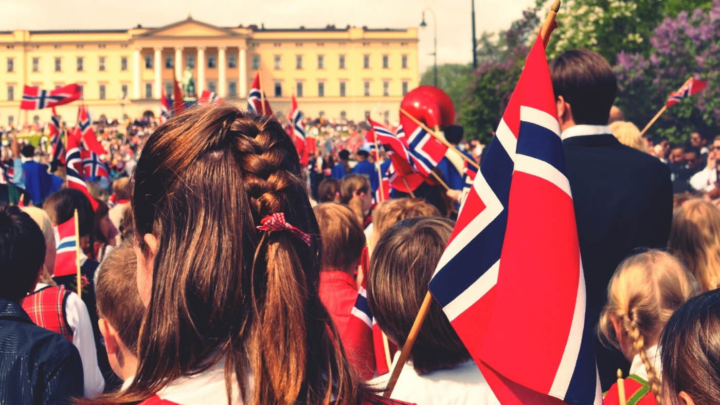 Mennesker står utenfor slottet med norske flagg på 17.mai, med fokus på jente med brunt hår med flette
