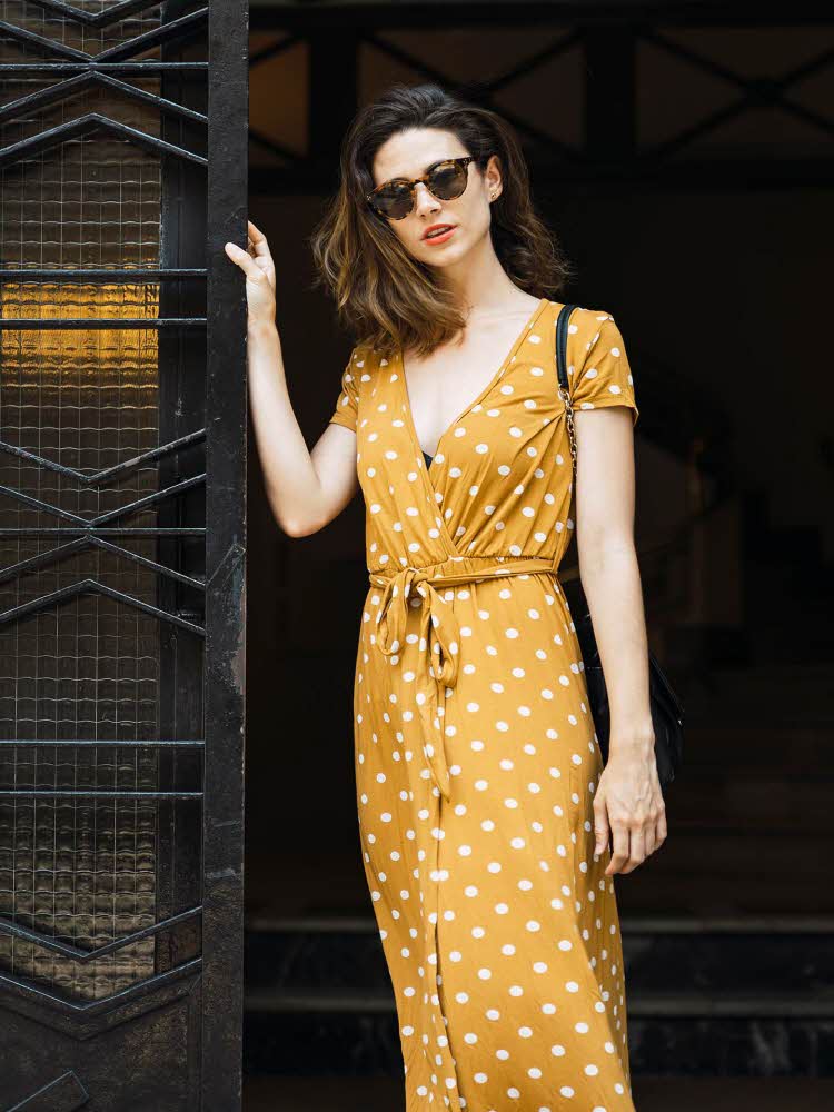 kvinne med gul, prikkete, lang, kortermet kjole og solbriller ved en port