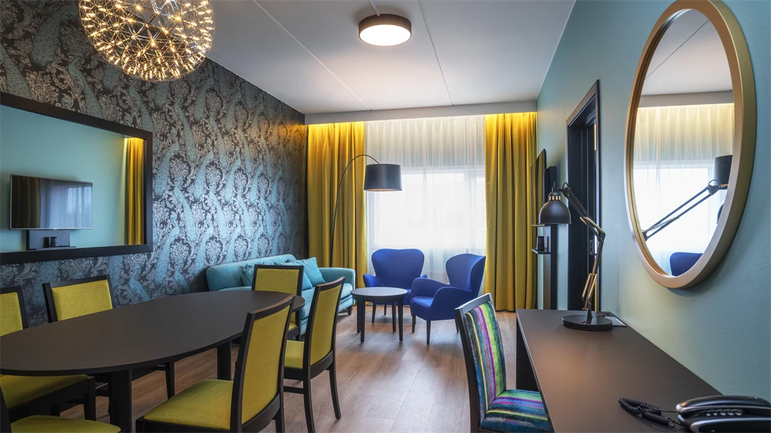 Tapetvegger med mønster, speil på veggen, vindu fra gulv til tak, gul gardin, turkis sofa, to blå stoler, parkettgulv, langt spisebord med gule stoler. Svart skrivebord med stol.