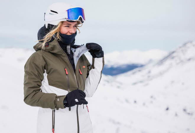 En kvinne i vinterklær, hjelm og skibriller står på en snøtopp