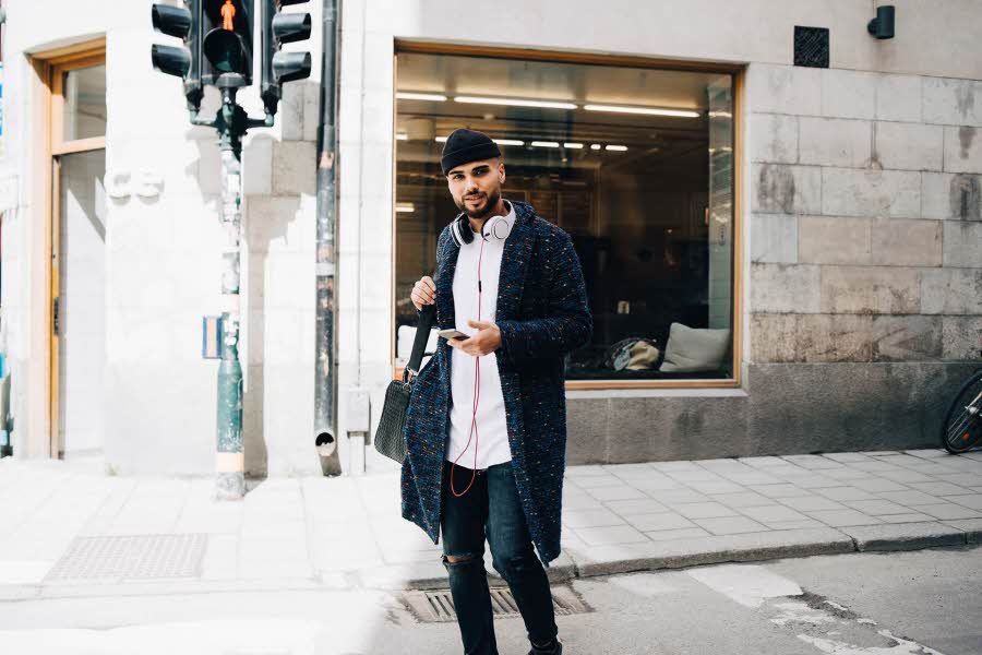 Mann med lang strikkejakke, lys skjorte, hullete skinny jeans, lue og veske med mobil i hånda, stående i urbant landskap