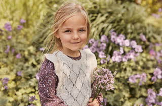En jente i burgunder kjole og krem vest står og holder en bukett med selvplukkede blomster.