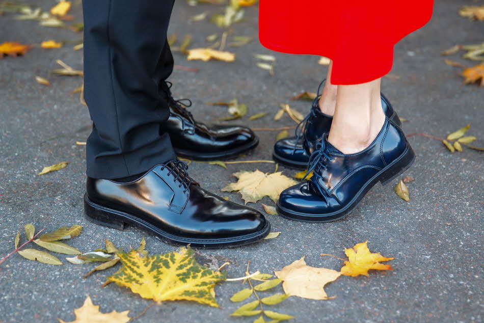Nærbilde av herreben med sort dressbukse med sorte skinnsko og dameben med rød kjole og sorte snøresko stående på asfalt med gule blader rundt
