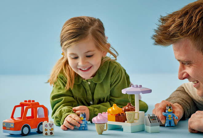 Et barn og en voksen leker med LEGO Duplo, inkludert biler, figurer og en iskrembod.