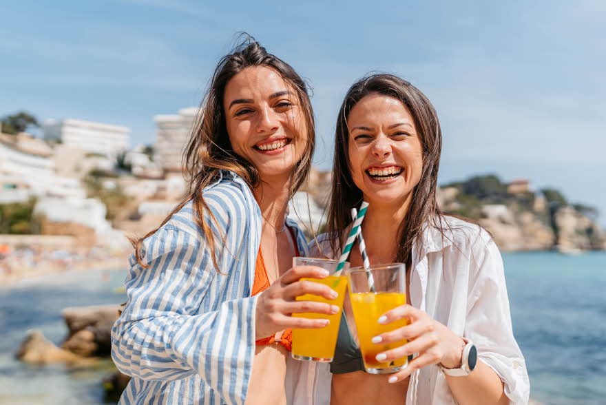 To damer med linskjorte står på strand med lemonade i hånden og smiler til kamera