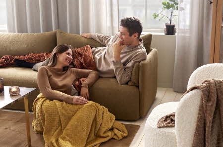 mann liggende på sofa med pledd og dame sittende på gulvet med pledd