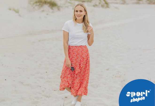 En kvinne i hvit t-skjorte og oransje blomstrete skjørt som går på en strand