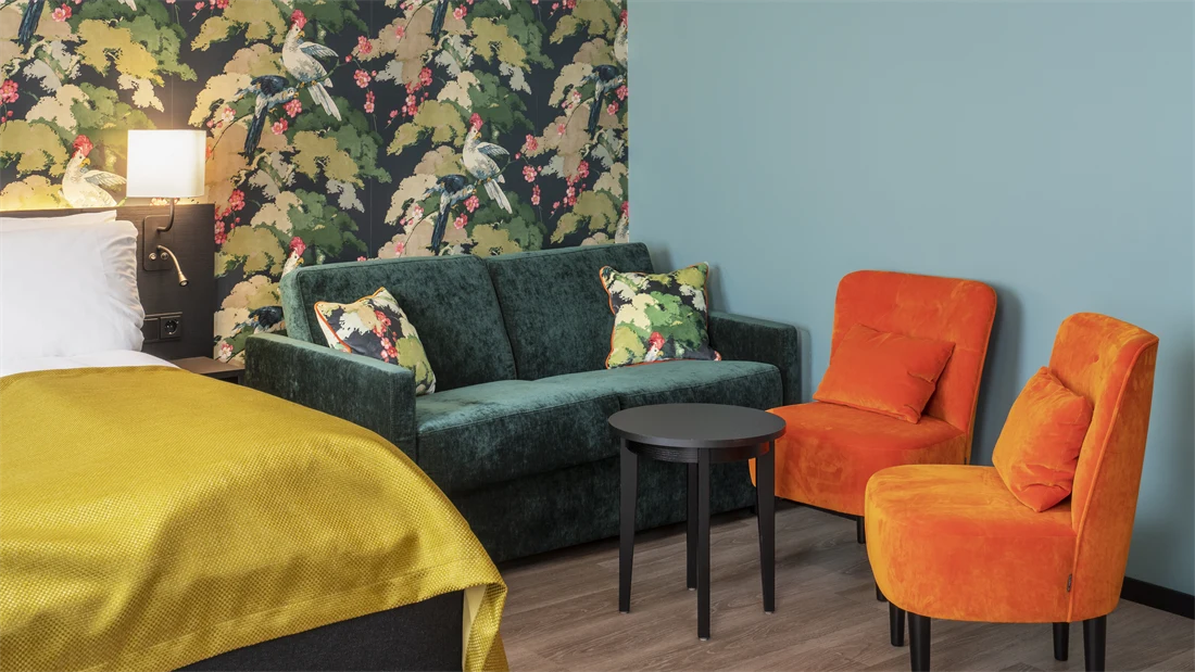 Seng med gult sengeteppe. Fargerik tapet med fugler og blomster, grønn sofa, oransje stoler og mørkt bord i business rom på Thon Hotel Bristol Bergen