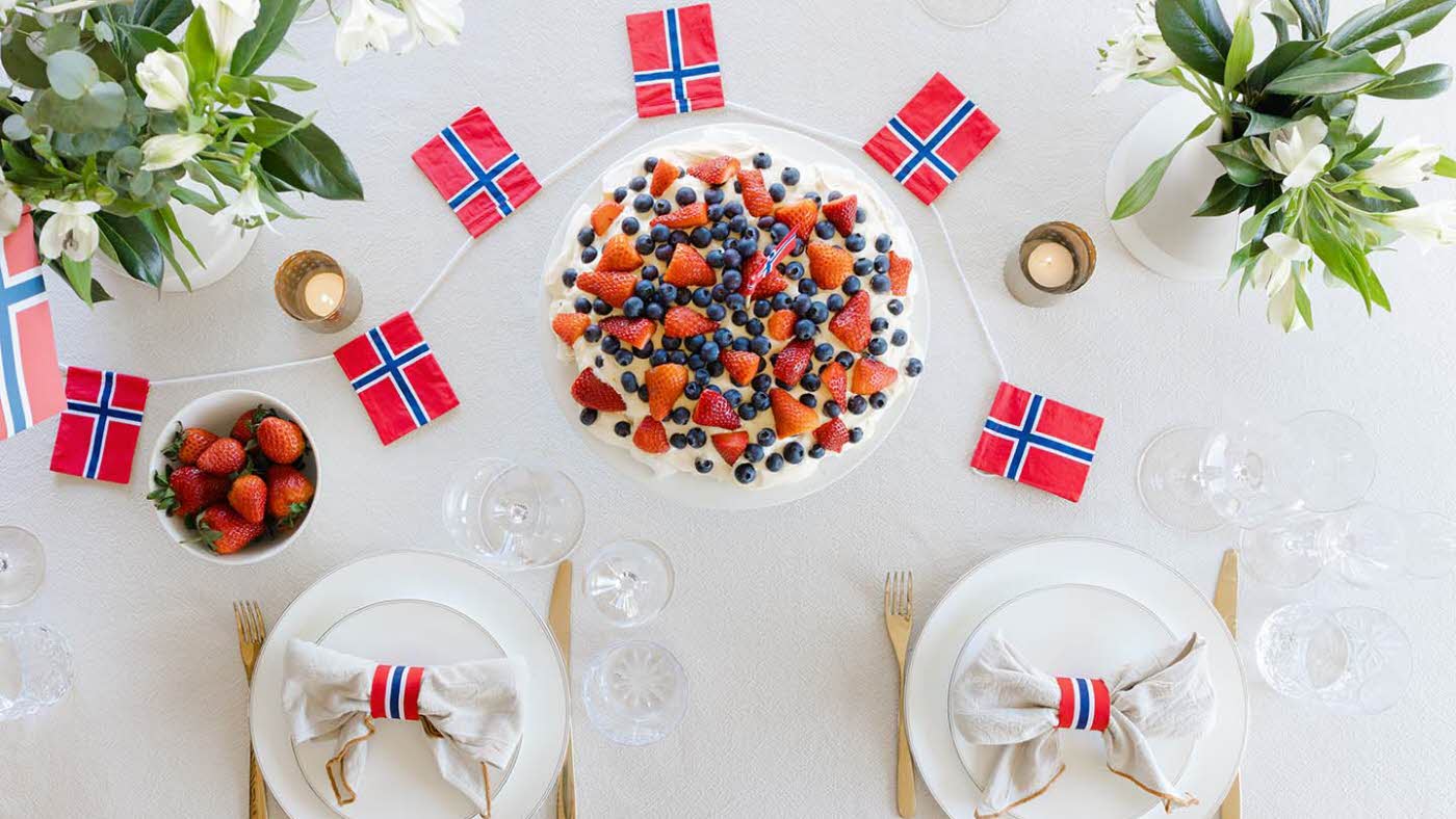 Fugleperspektiv ned på dekket bord, med hvit duk, fine tallerkener, kake og flagg i rødt, hvitt og blått, friske vårblomster. Foto til artikkel om borddekking på 17. mai.