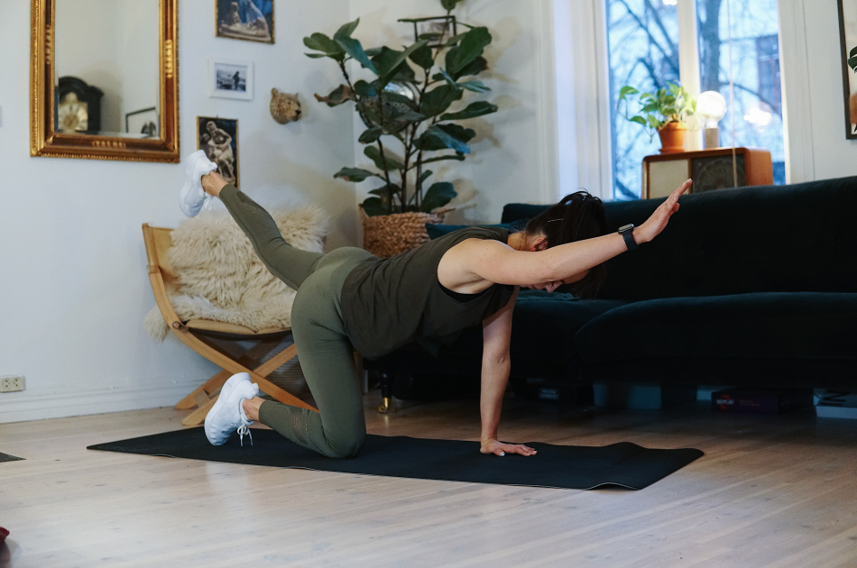 treningsekspert Anna Johansen trener på matte på gulvet, mørkegrønn sofa i bakgrunnen, lys stol med pels oppi, str grønn plante, bilder på veggen