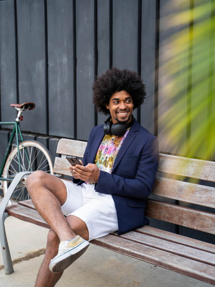 Mann med blå blazerjakke, mønstret t-skjorte og hvit shorts med headset rundt halsen sitter på en benk utendørs og smiler med en palme i forgrunnen