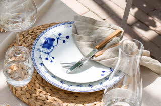 Et bordoppsett med hvite og blå tallerkener, en flettet bordbrikke, en beige linserviett og bestikk.