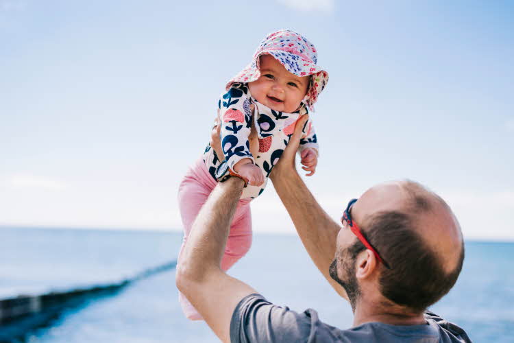 Mann holder opp baby som smiler i mønstrede klær og solhatt