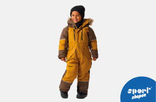 Gutt står i gul og brun vinterdress med pelskant på hetten og svarte støvler.