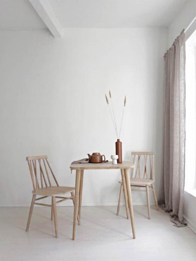 Hvitt rom med et kjøkkenbord og to pinnestoler i lyst tre. Brun tekanne og vase med strå i står på bordet. Duse sandfargede gardiner henger foran et vindu til høyre. 