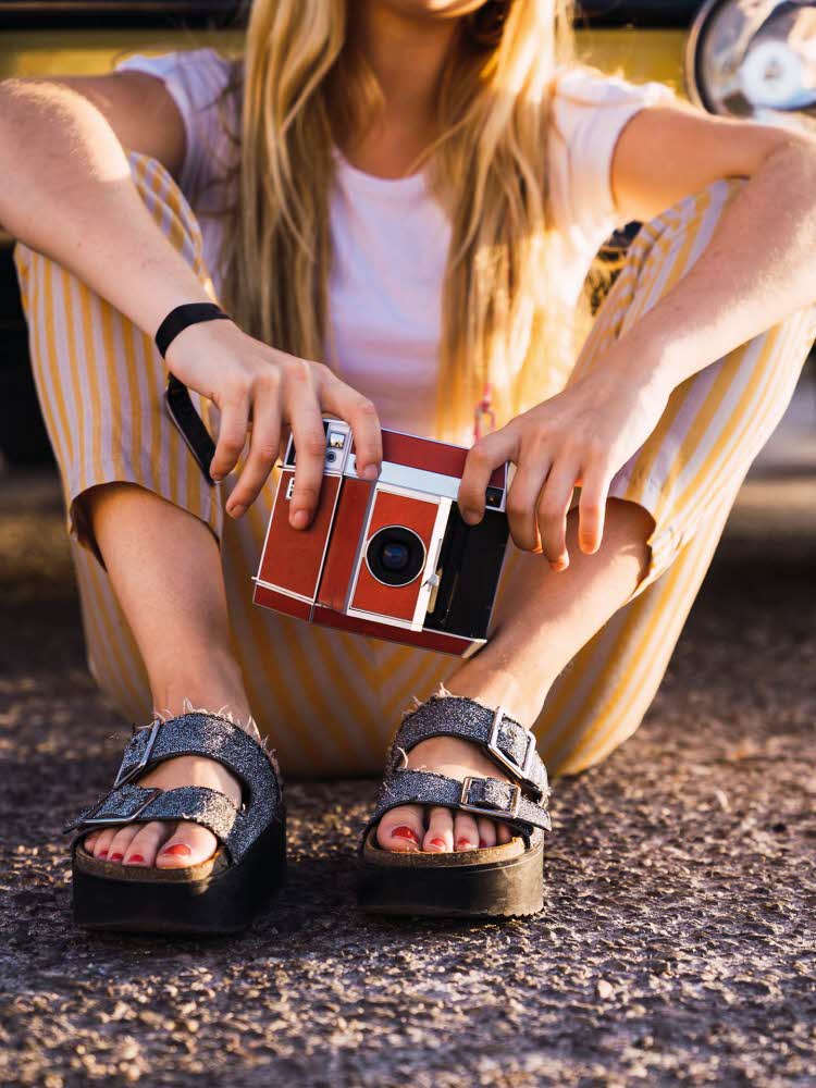 Kvinne med platåsandaler med dobbel reim som sitter på bakken og holder et kamera. Illustrasjonsbilde til artikkel om vårmote for sko.