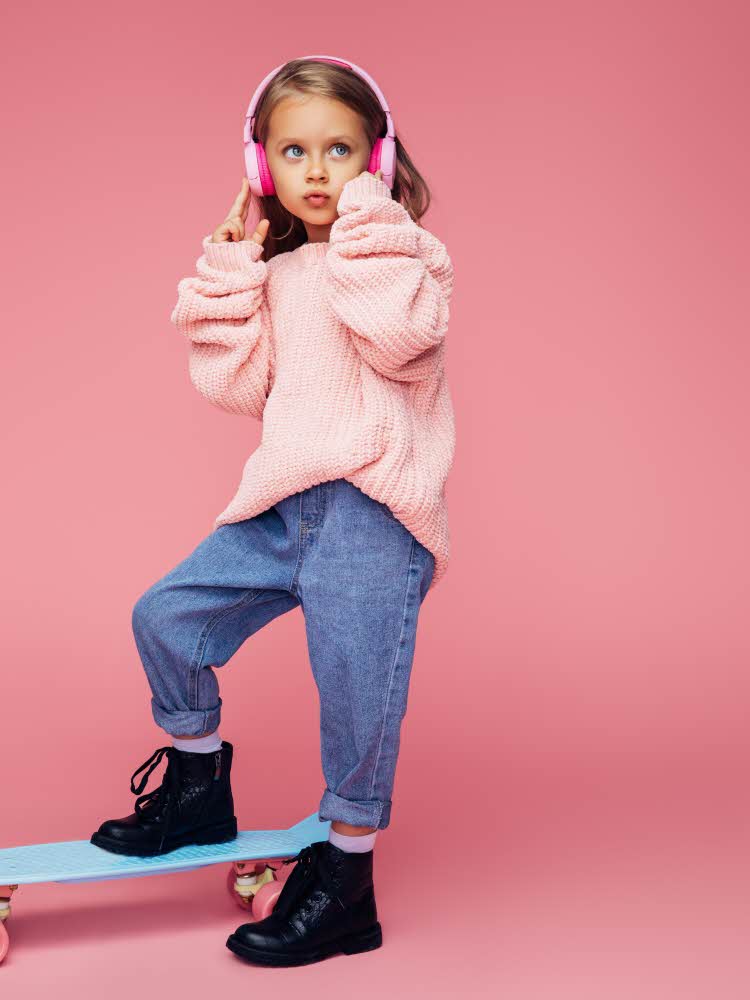 poserende jente med rosa genser, jeans, støvler med headsett og skateboard poserende jente med beige frakk, sort kjole, hvite sokker og støvler