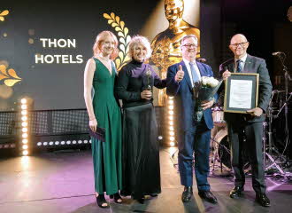 To damer og to menn i pene klær står på scene under prisutdeling, med blomster og diplom, stor skjerm bak seg der det står Thon Hotels i gullskrift.