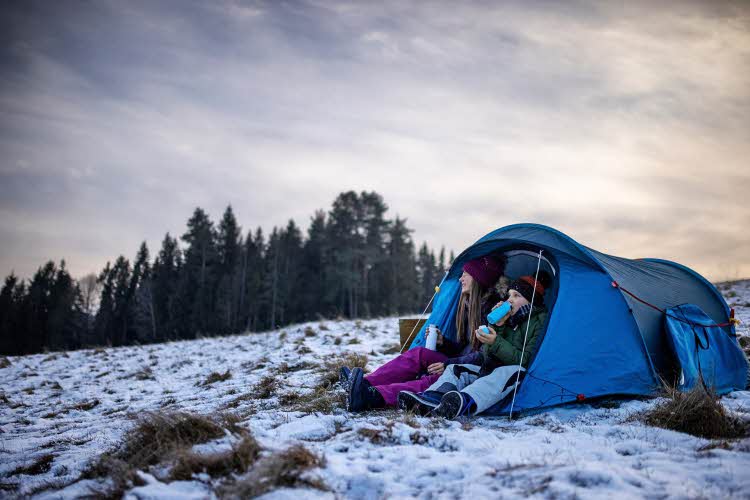 To unger som sitter i en teltåpning i delvis snølandskap