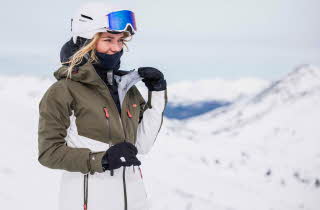 En kvinne i vinterklær, hjelm og skibriller står på en snøtopp