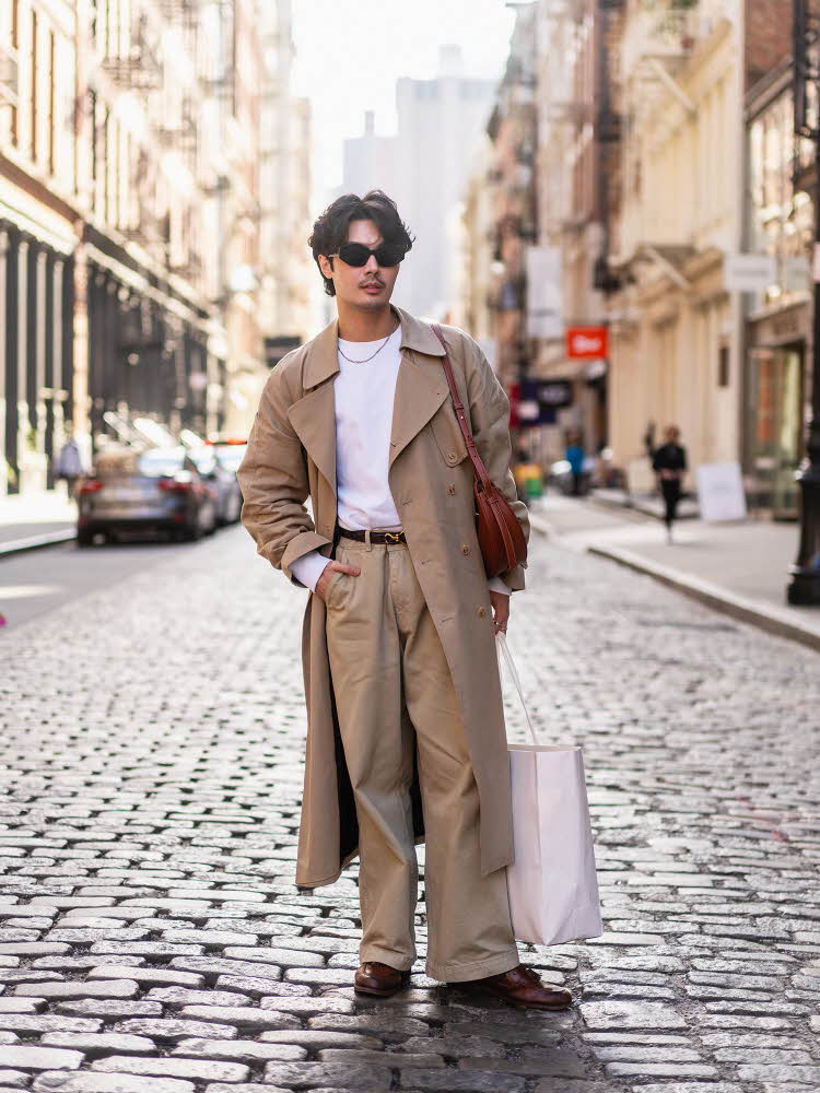 Mann med beige vide dressbukser, hvit t-skjorte, beige trenchcoat og rød veske poserer på gaten