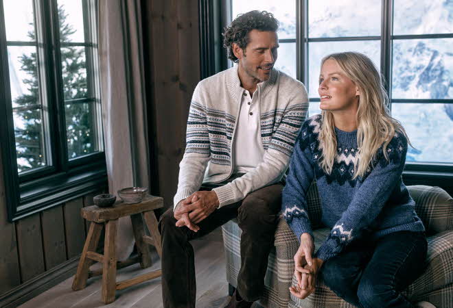 En mann og en dame i ullklær sitter inne med utsikt mot snødekte fjell.