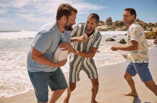 Tre menn i tskjorte og shorts spiller rugby på en strand ved vannet