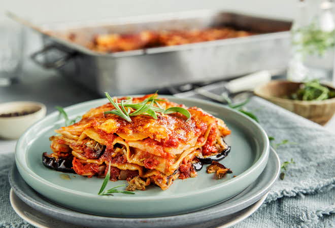 Et stykke lasagne på en blå tallerken, med mer lasagne i en form i bakgrunnen.