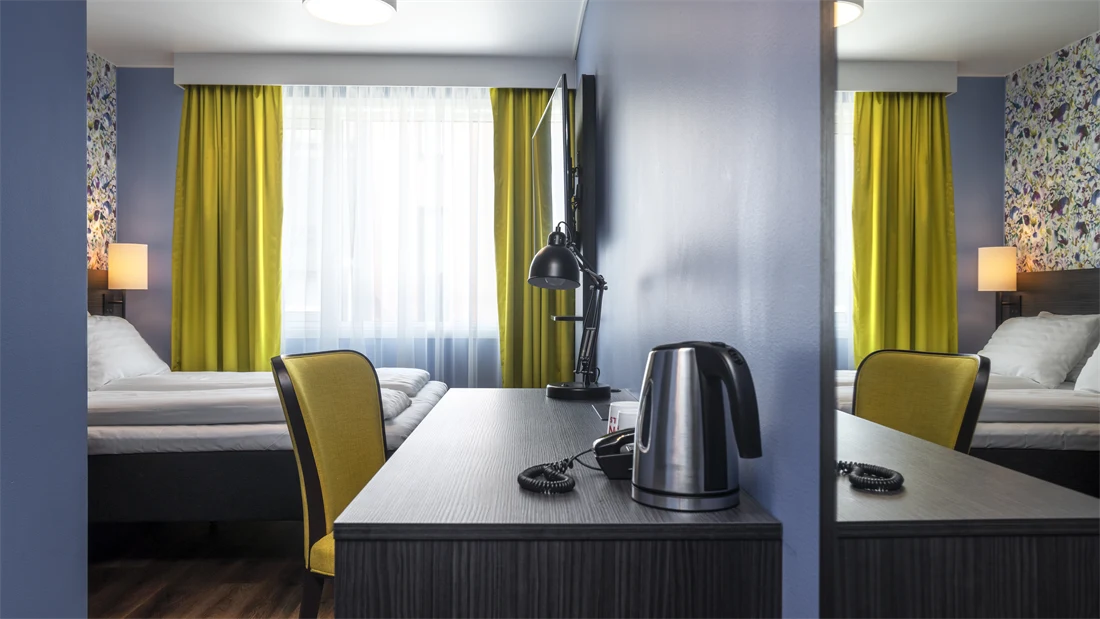 Tapetvegg, dobbeltseng, skrivebord, blå vegg, vindu, gule gardiner.