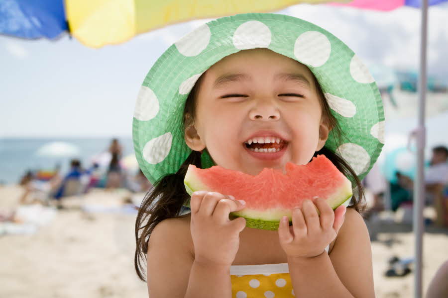 Jente med hatt spiser melon på stranda