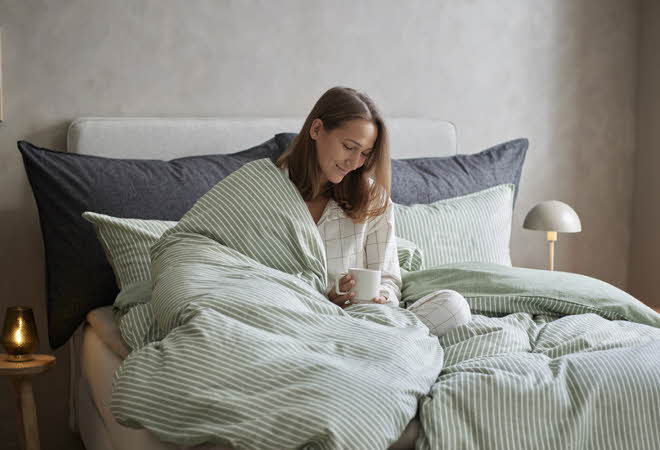 En kvinne som sitter i sengen med et grønt flanellsengesett og holder en kaffekopp