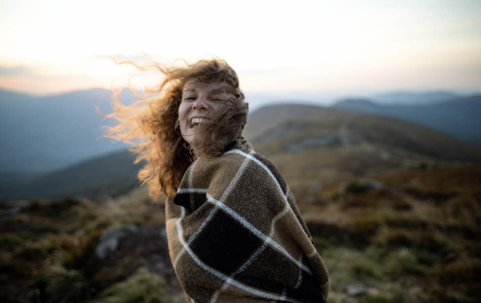Dame står i vind og solnedgang på en fjelltopp og holder et stort skotskrutet sjal rundt kroppen.