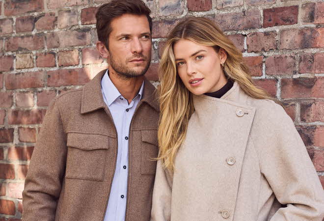 En mann i brun jakke og en kvinne i beige jakke lener seg mot en mursteinsvegg.