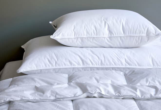 Hvite puter og dyne stablet på en seng mot en grå vegg.