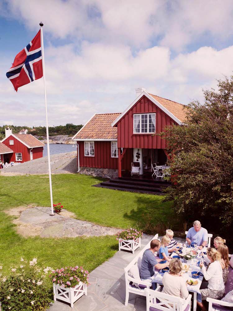 Oversiktsbilde over hage ved sjøen med familie som spiser ved et bord og flaggstang med norsk flagg. Foto til artikkel om 17. mai-dekorasjoner.