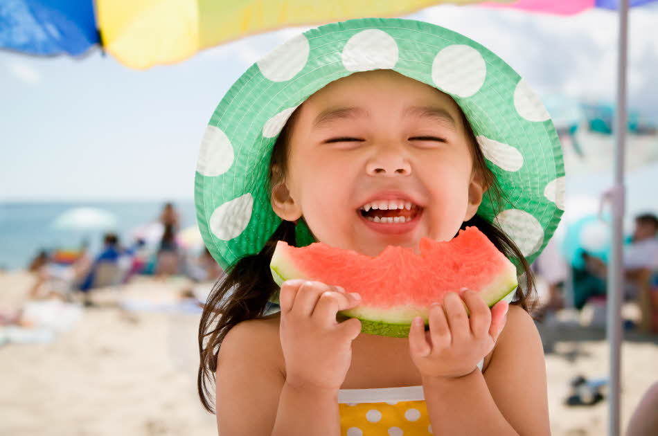 Jente med hatt spiser melon på stranda