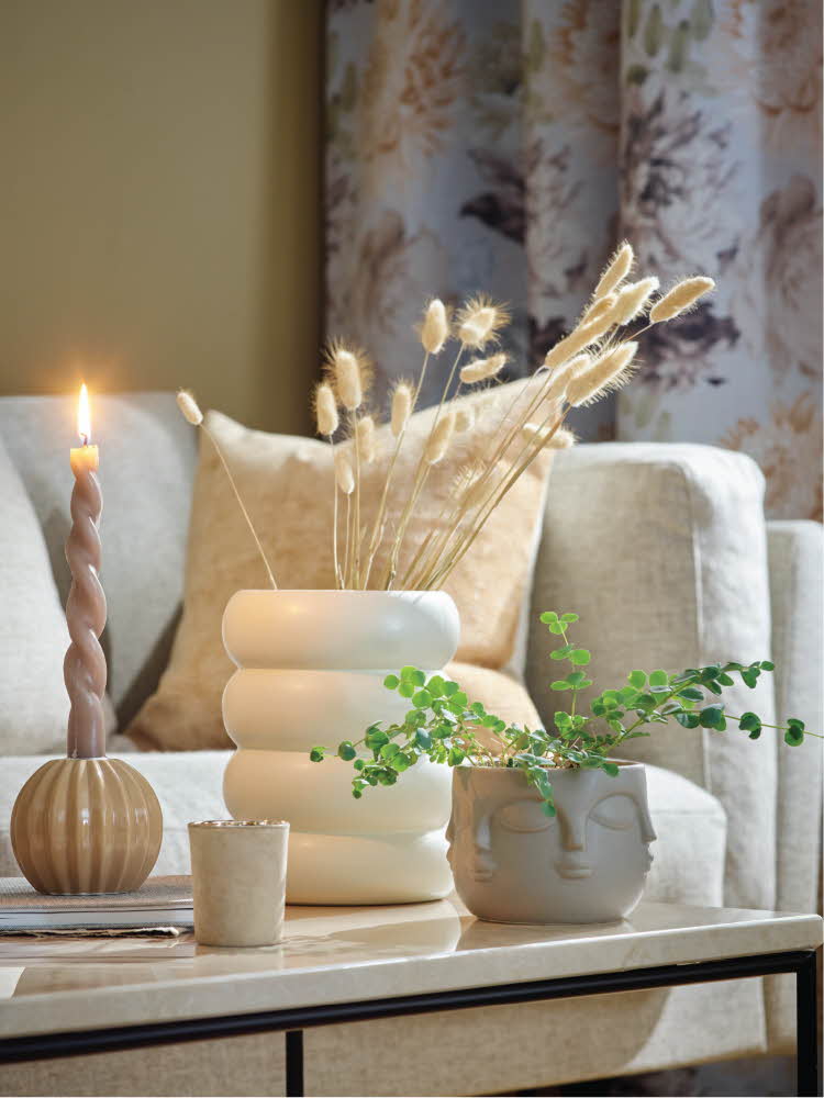 Lys sofa og et bord dekket med en vase med strå, grønn plante og tent lys