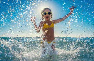 En liten jente i bikini og solbriller som løper gjennom vannet, slik at det spruter rundt henne