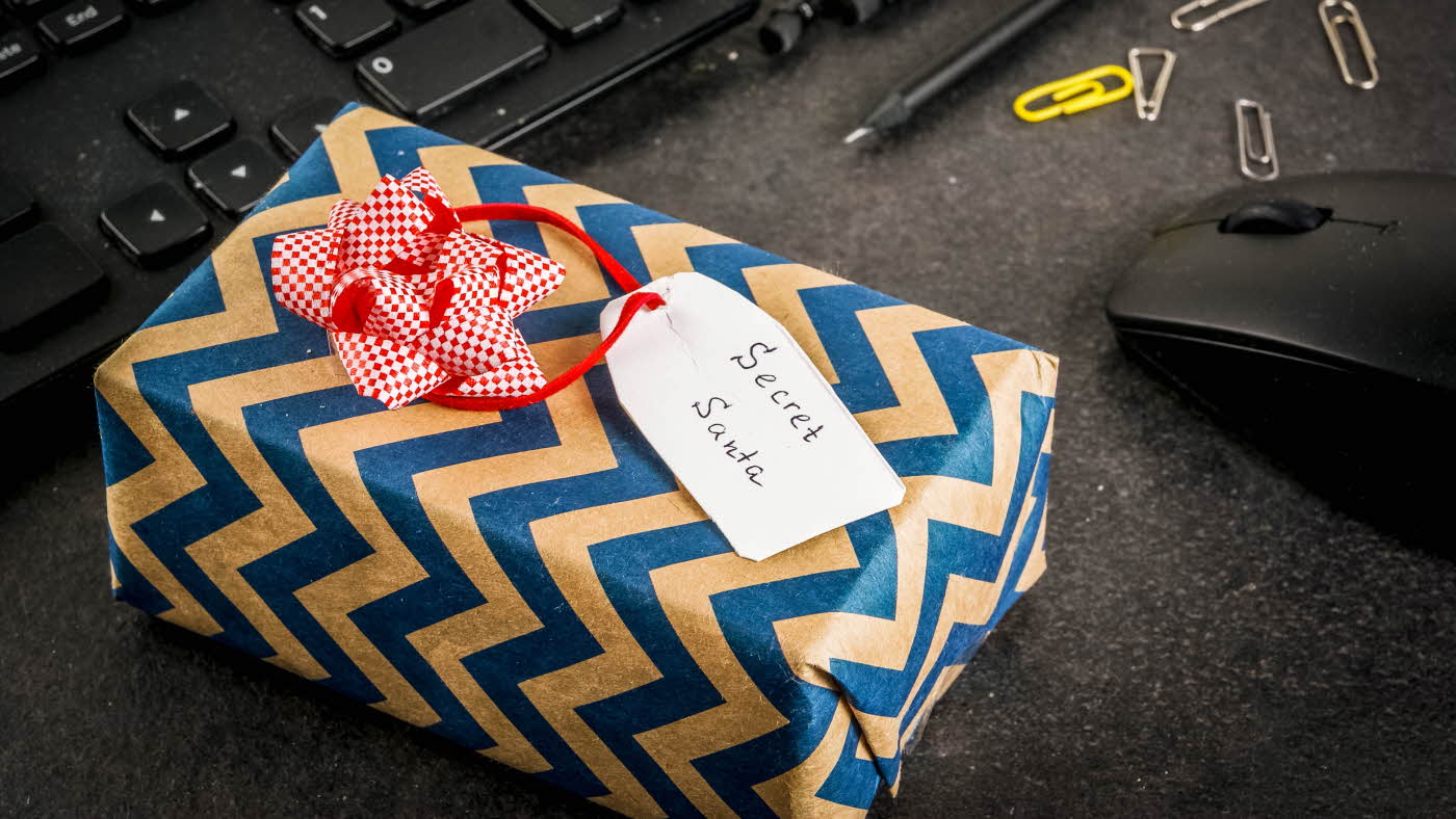 Innpakket, firkantet gave med påskriften "Secret Santa" på pult ved mus, penn, binders og tastatur.