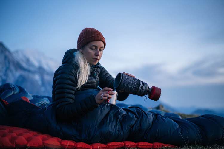 Kvinne som sitter på liggeunderlag inni sovepose ute i snøen og heller varm drikke fra termos