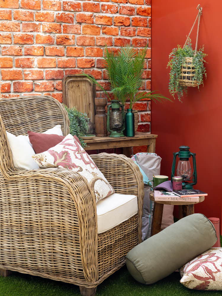 Kurvstol med puter, lite trebord med bøker og lykt på, plante på veggen i bakgrunnen, puter på gulvet foran stol