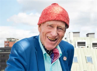 Nærbilde av Olav Thon, med signatur-luen, med en fin utsikt av Oslo bak