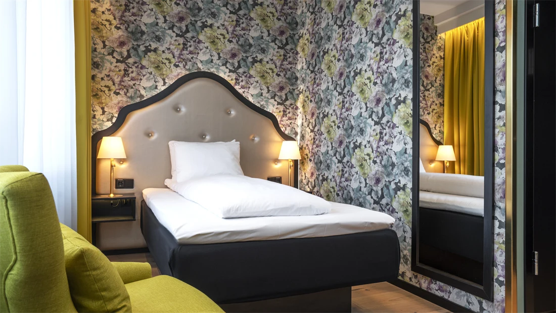 Gul stol, queen seng med sengegavl, blomstrete tapet og stort vegghengt speil i standard rom queen på Thon Hotel Cecil.