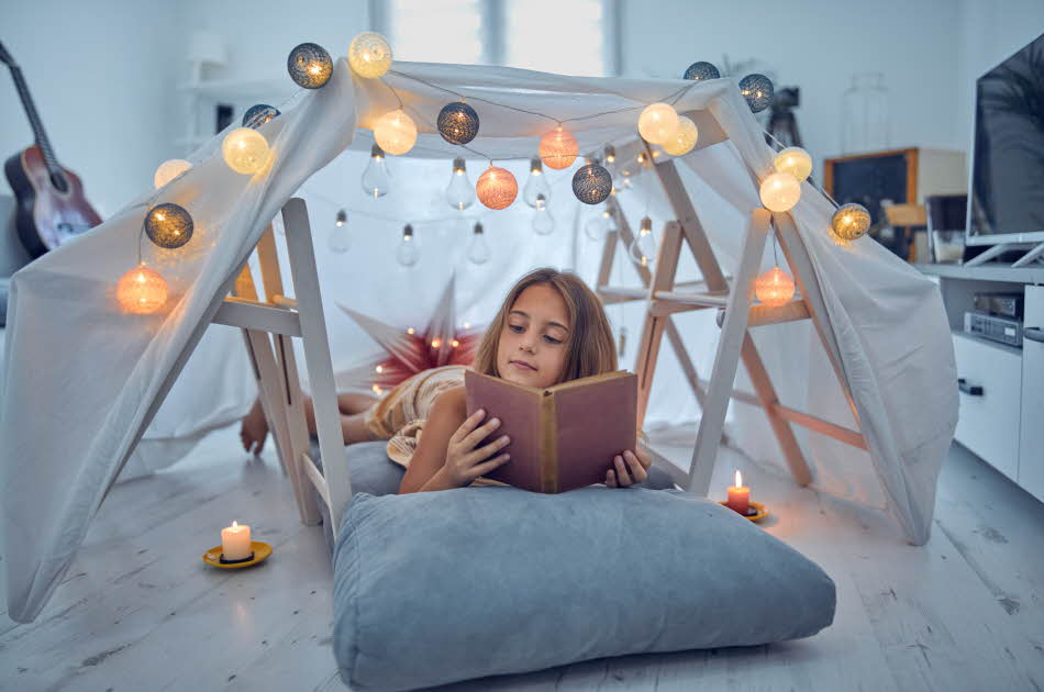 Jente som leser bok på gulvet i sitt hjemmelagde telt i stua